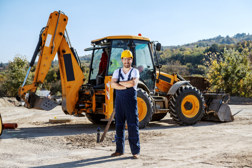 A Construction Worker Wearing A Yellow Hard Hat And Blue Overalls Standing Confidently With Arms Crossed In Front Of A Yellow Excavator On A Sunny Construction Site With Trees And Hills In The Background